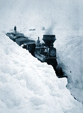 Photo of a train stuck in the snow. The snowbank walls are as tall as teh roofs of the engine and cars.